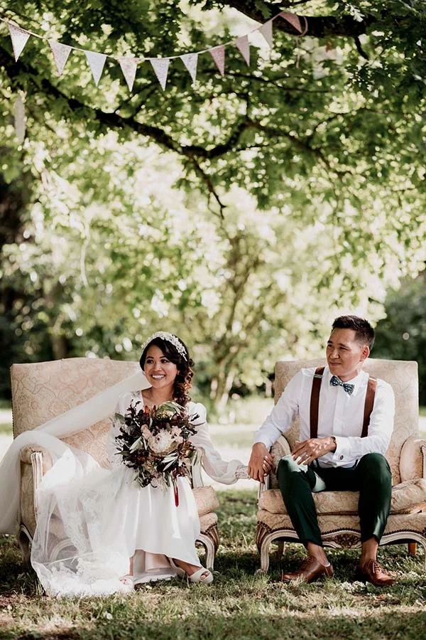 Jeunes mariés en tenue bohème assis dans des fauteuils vintage sous des arbres, ambiance douce et naturelle.