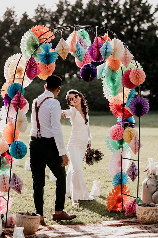 Couple de mariés devant une arche colorée en éventails de papier pour un mariage bohème et artisanal.