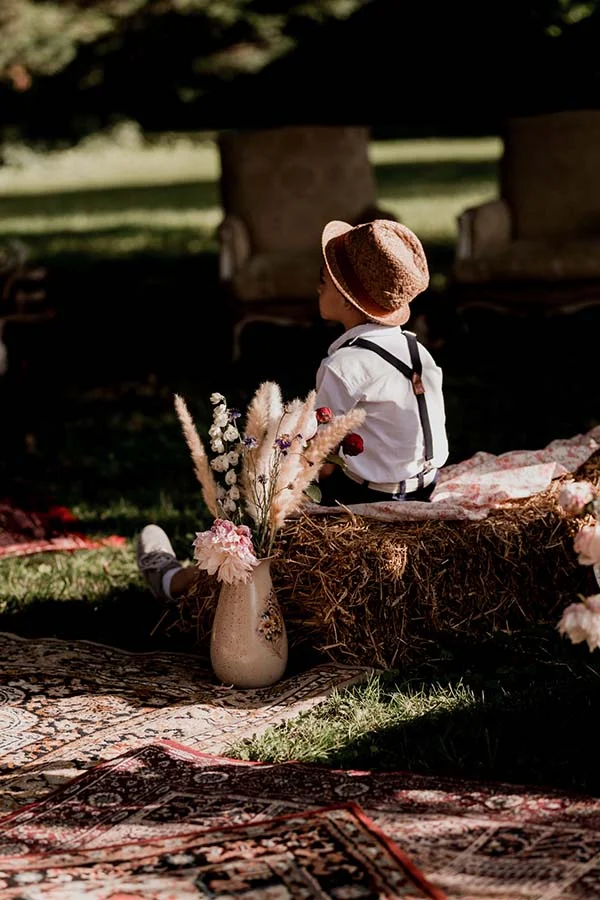 Petit garçon au style bohème avec chapeau et bretelles, assis sur une botte de paille décorée de fleurs séchées.