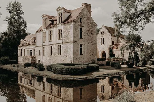 Manoir ancien en pierre claire au bord de l'eau, reflété dans un étang sous une lumière douce et romantique.