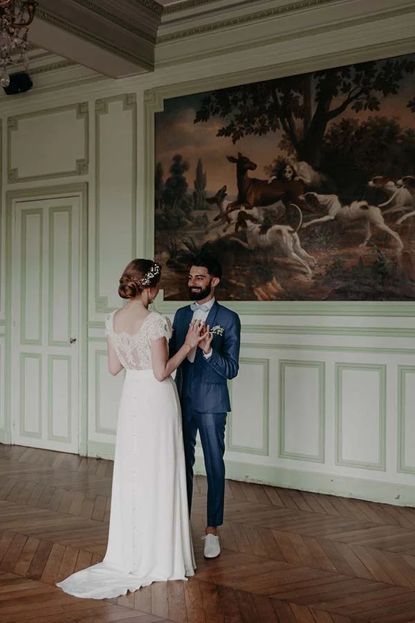 Jeunes mariés face à face dans un salon classique devant une grande tapisserie, ambiance romantique et élégante.