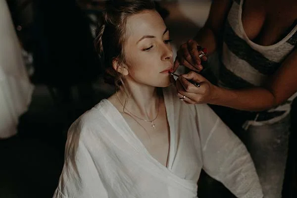 Une maquilleuse applique du rouge à lèvres à une mariée en peignoir blanc dans une ambiance douce et intimiste.