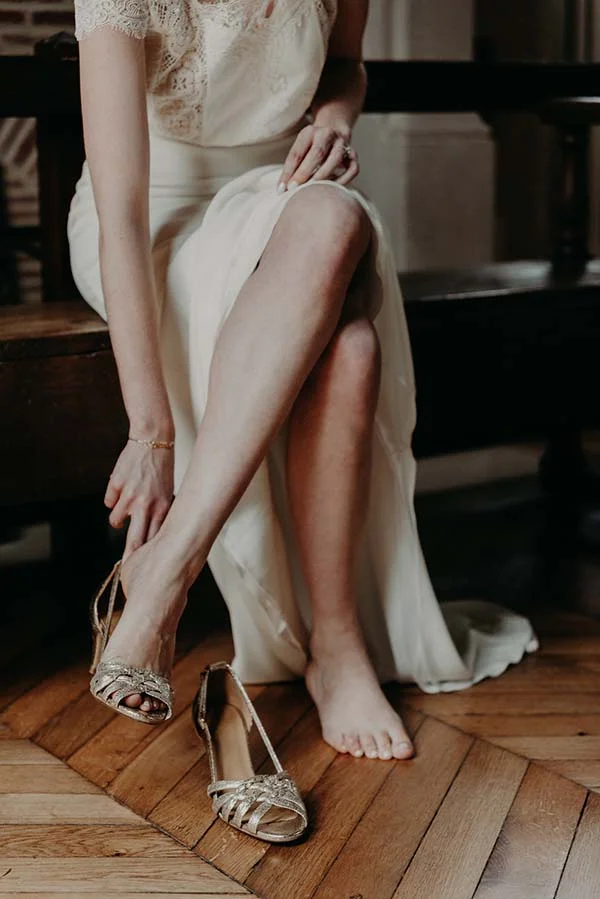 Une mariée en robe blanche et dentelle enfile ses sandales dorées sur un parquet en chevrons.