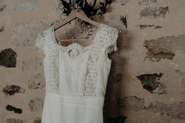 Robe de mariée blanche en dentelle suspendue devant un mur en pierres rustiques.