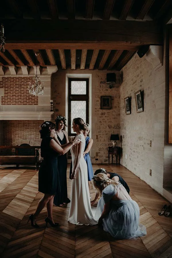 Une mariée en robe blanche se fait aider par ses demoiselles d'honneur dans un salon rustique et chaleureux.