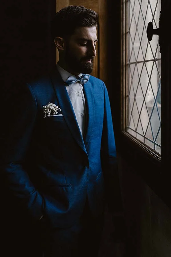 Homme barbu en costume bleu royal et nœud papillon, regardant pensivement par une fenêtre à carreaux.
