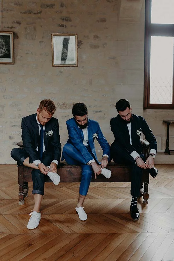 Trois hommes en costumes bleu marine et bleu roi s'apprêtent, assis sur un banc dans un intérieur aux murs de pierre.