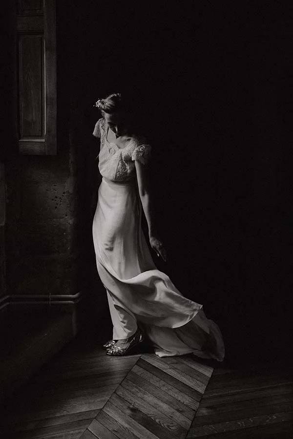 Femme en robe de mariée blanche et chaussures scintillantes, pose élégante dans un clair-obscur dramatique et intemporel.