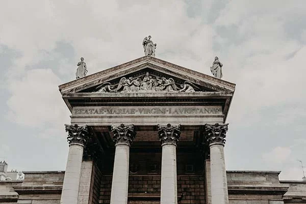 Façade néoclassique en pierre claire d'une église avec colonnes corinthiennes et fronton sculpté sous un ciel nuageux.