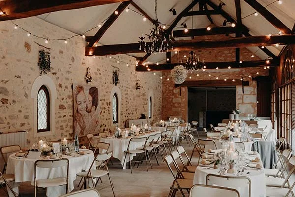 Salle de réception rustique et chaleureuse avec tables dressées, poutres en bois et guirlandes lumineuses.