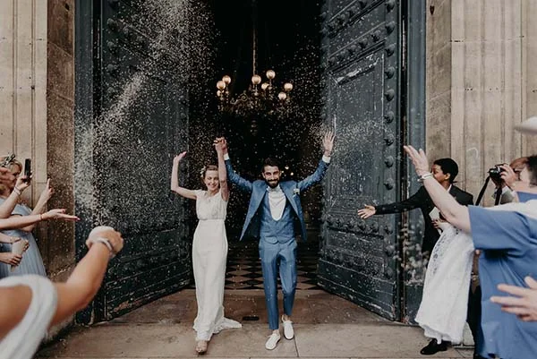 Jeunes mariés joyeux sortant d'une église sous une pluie de confettis, portant une robe blanche et un costume bleu.