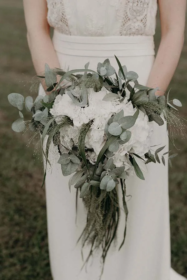 Mariée en robe blanche tenant un bouquet composé de fleurs blanches et de feuillage d'eucalyptus vert gris.