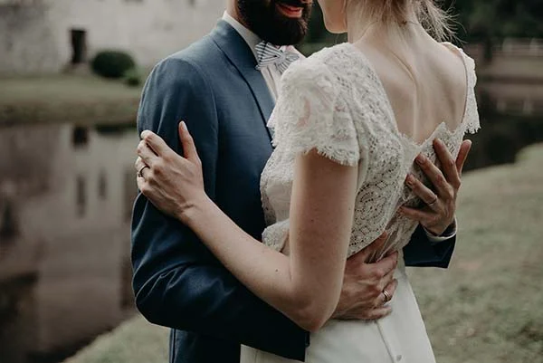 Jeunes mariés s'enlaçant tendrement, elle en robe de dentelle blanche et lui en costume bleu marine.