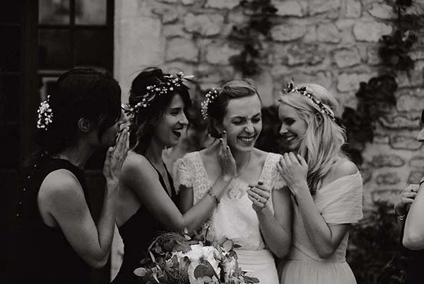 Quatre femmes rient ensemble en noir et blanc, dont une mariée et ses demoiselles d'honneur couronnées de fleurs.