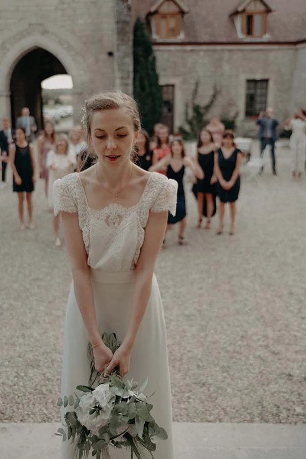 Une mariée en robe blanche élégante tenant un bouquet, entourée de ses invitées devant un château en pierre.