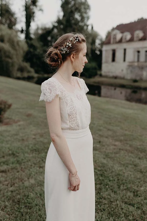 Mariée de profil en robe blanche et dentelle, portant un accessoire floral, dans un jardin romantique devant un château.