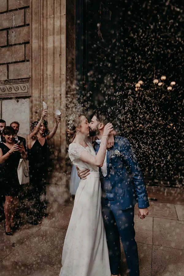 Jeunes mariés s'embrassant sous une pluie de confettis, elle en robe blanche et lui en costume bleu, ambiance festive.