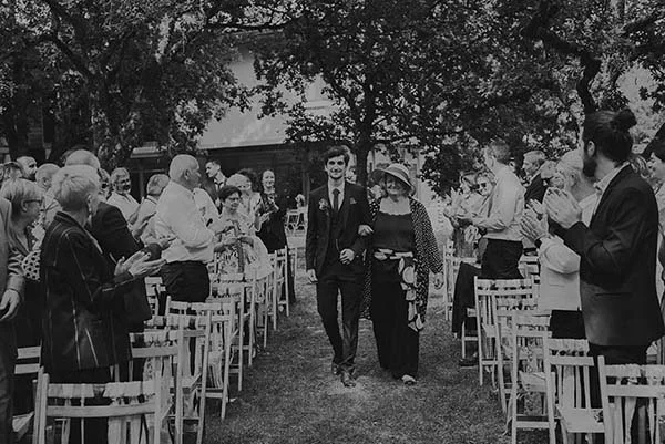 Marié et sa mère remontent l'allée d'une cérémonie en plein air, entourés d'invités, dans un style bohème en noir et blanc.