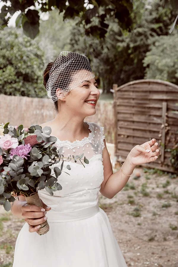 Mariée souriante en robe blanche avec voilette et bouquet champêtre, dans un cadre extérieur naturel et lumineux.