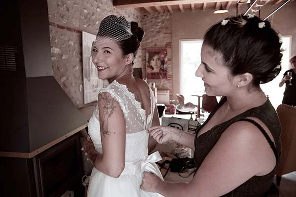Une femme aide la mariée à ajuster sa robe blanche dans un intérieur rustique aux tons sépia.