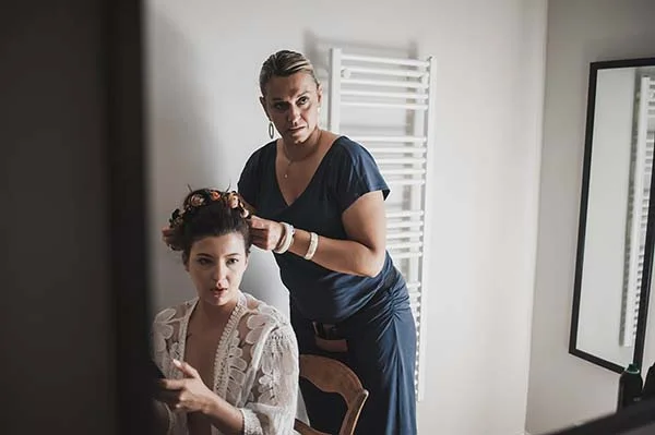 Une coiffeuse ajuste les accessoires de cheveux d'une mariée en robe blanche dans une ambiance lumineuse et sereine.