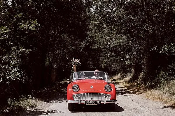 Jeunes mariés souriants dans une décapotable rouge vintage sur un chemin ombragé et champêtre.