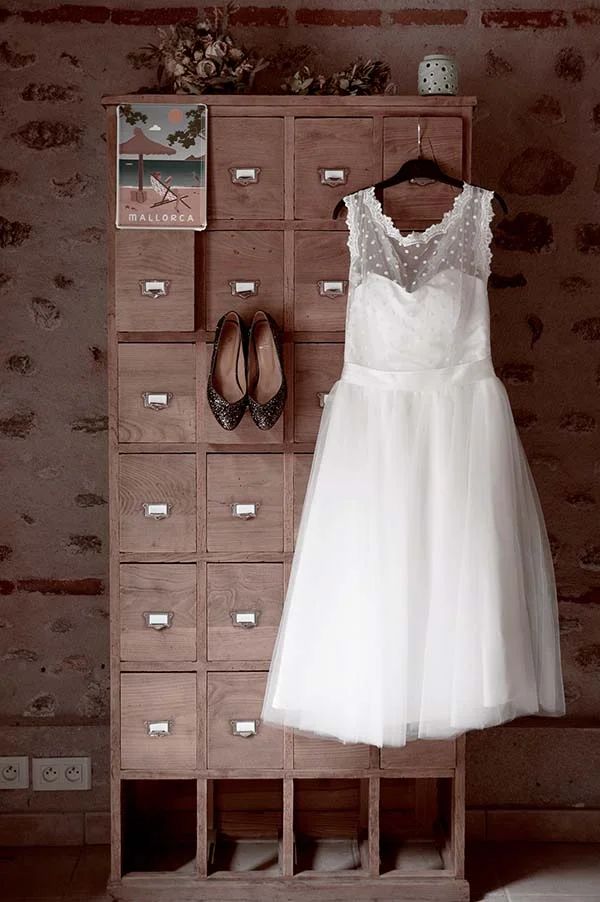 Robe de mariée blanche et escarpins pailletés suspendus à un meuble en bois rustique devant un mur en pierres.