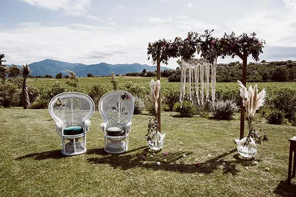 Arche fleurie et fauteuils en osier blanc pour une cérémonie de mariage champêtre en plein air face aux montagnes.