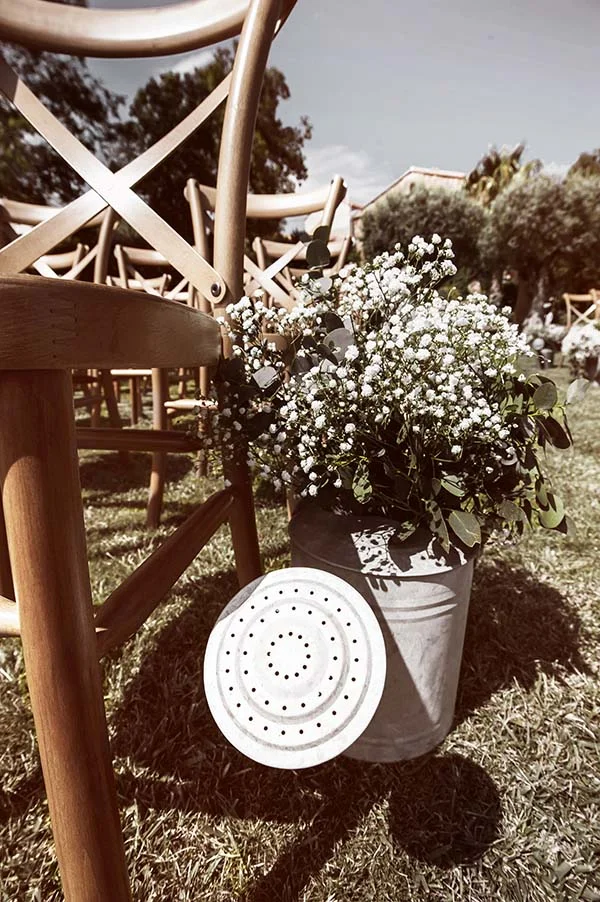 Bouquet de gypsophile blanc dans un arrosoir vintage au pied de chaises en bois, ambiance champêtre et lumineuse.