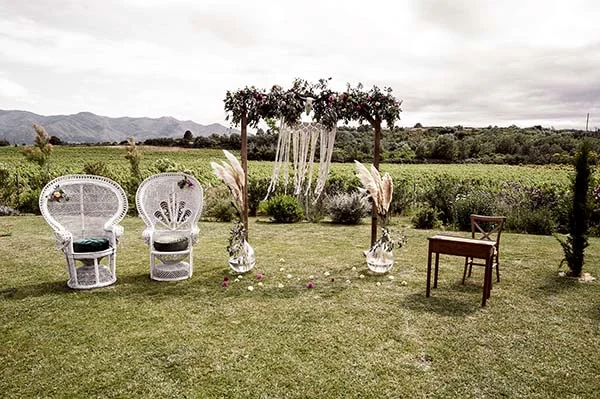 Arche fleurie et fauteuils en osier blanc pour une cérémonie champêtre en plein air face aux montagnes.