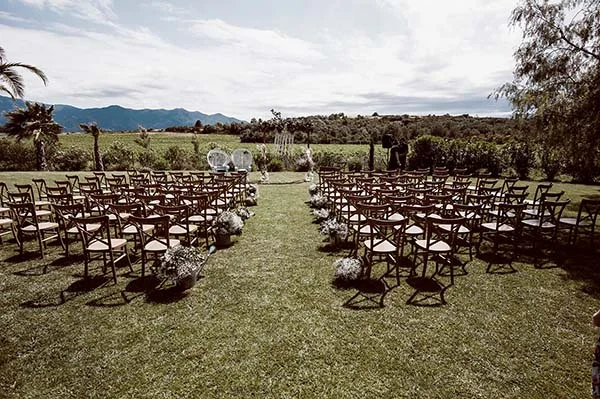 Installation de chaises en bois pour une cérémonie de mariage champêtre en plein air sous un ciel lumineux.