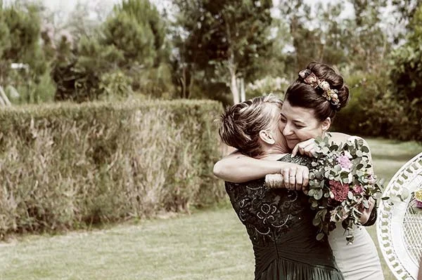 Deux femmes s'enlaçant tendrement dans un jardin champêtre, l'une en robe blanche et l'autre en robe vert foncé.
