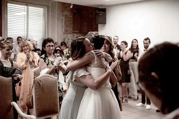 Deux mariées s'enlaçant avec émotion devant leurs invités lors d'une réception lumineuse et chaleureuse.