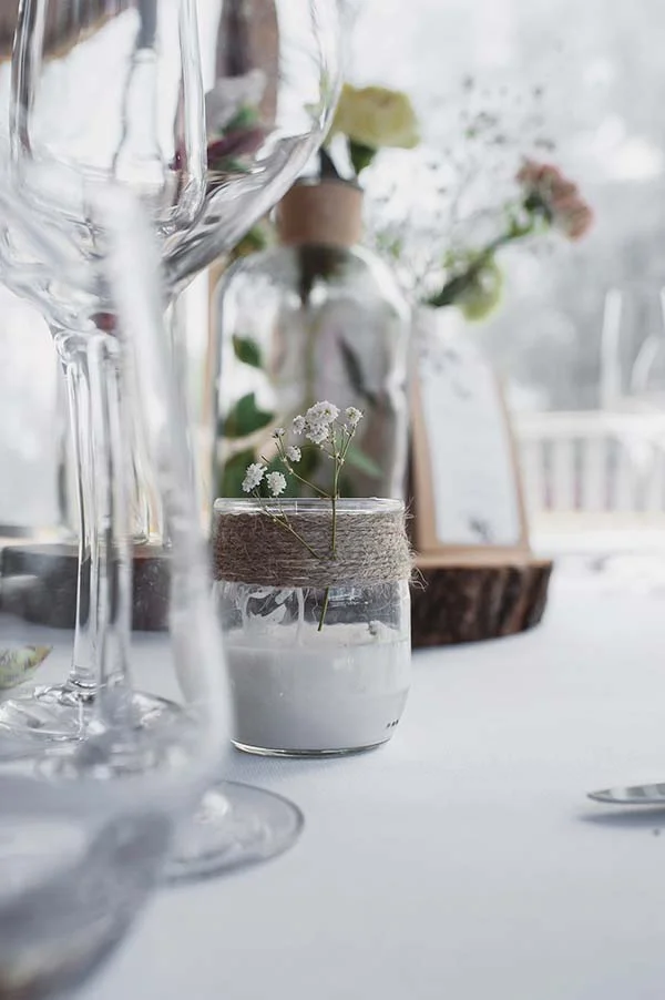Décoration de table champêtre avec un petit vase en verre entouré de ficelle et des fleurs blanches délicates.