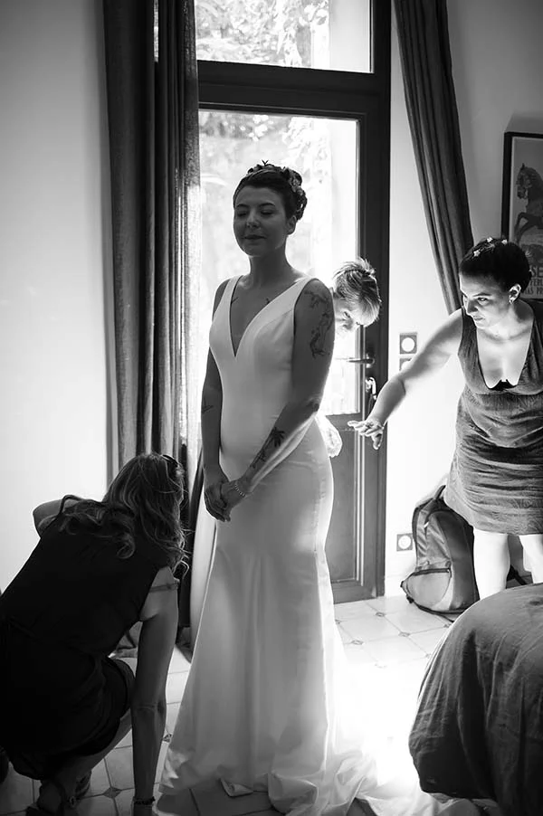 Photo en noir et blanc d'une mariée en robe blanche se faisant aider pour sa tenue dans une ambiance intime.