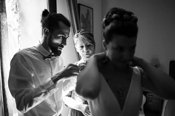 Photo noir et blanc d'un marié et d'une femme aidant la mariée à s'habiller dans une ambiance intime.