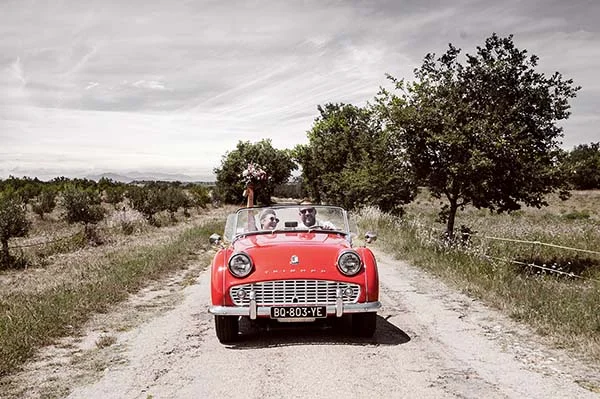 Couple souriant dans une décapotable rouge vintage sur un chemin de campagne, ambiance champêtre et lumineuse.