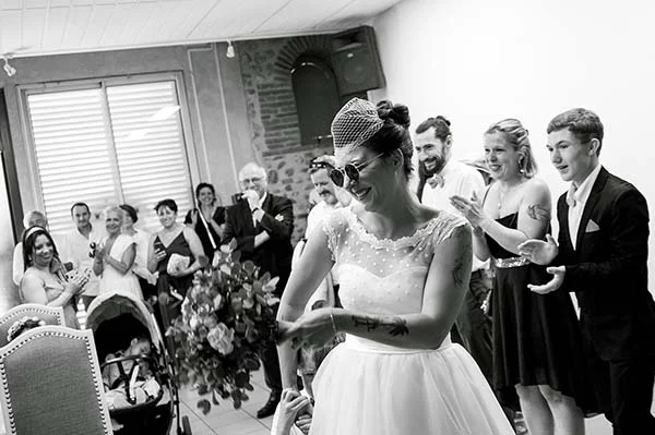 Mariée souriante en robe blanche et lunettes de soleil, entourée d'invités joyeux, dans un cliché noir et blanc.