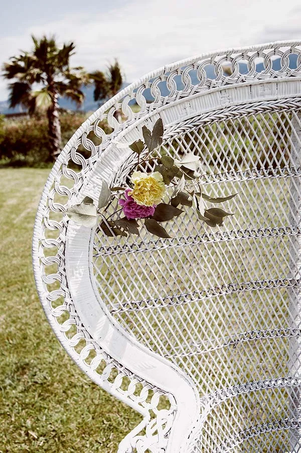 Fauteuil en osier blanc décoré de fleurs jaunes et roses dans un jardin champêtre avec des palmiers en arrière-plan.