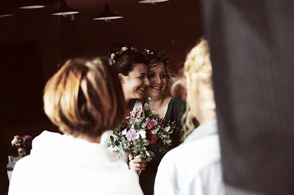 Une mariée souriante tenant un bouquet champêtre, entourée de ses proches dans une ambiance chaleureuse et intimiste.