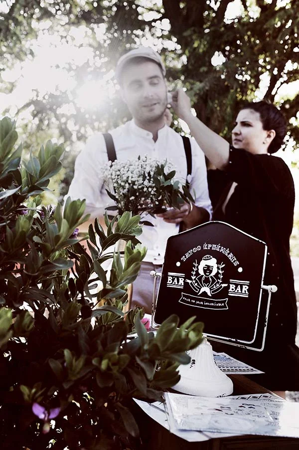 Un homme en tenue champêtre et une femme près d'un panneau de tatouages éphémères dans un jardin lumineux.