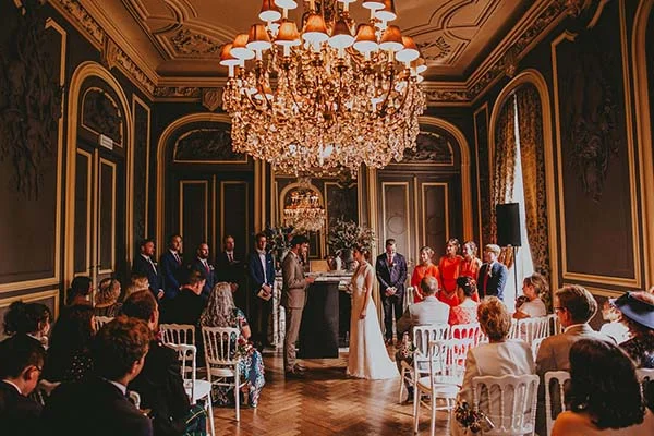Cérémonie de mariage élégante dans un salon bourgeois aux tons sombres, sous un immense lustre doré et scintillant.