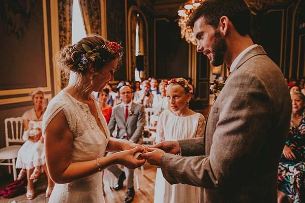 Jeune couple s'échangeant les alliances lors d'une cérémonie chaleureuse dans un intérieur élégant et coloré.