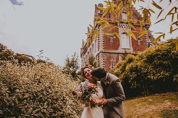 Jeunes mariés s'embrassant tendrement devant un château belge, entourés de fleurs colorées et de végétation luxuriante.