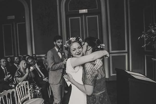 Photo noir et blanc d'une mariée à la couronne de fleurs embrassant une invitée, entourées de convives élégants.