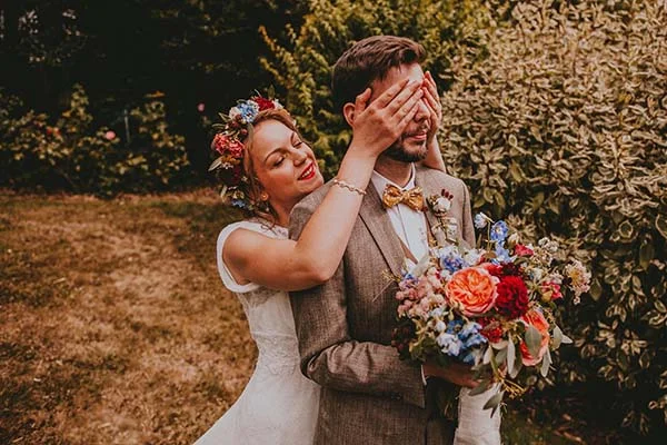 Une mariée à la couronne de fleurs couvre les yeux de son époux en costume beige dans un jardin champêtre coloré.