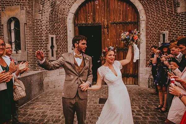 Jeunes mariés joyeux devant une porte en bois, elle en robe blanche et couronne de fleurs, lui en costume marron.