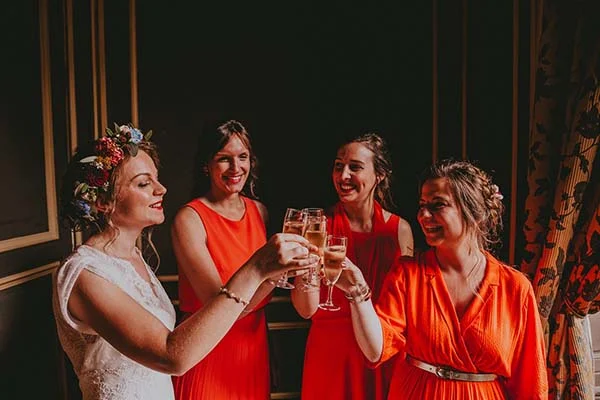 Une mariée et ses demoiselles d'honneur en robes orange trinquant avec des coupes de champagne dans une ambiance festive.