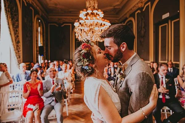 Jeunes mariés s'embrassant dans un salon doré, la mariée portant des fleurs colorées dans les cheveux.