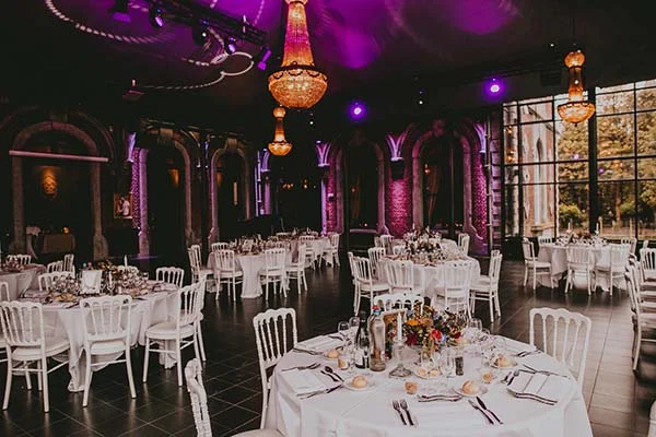Salle de réception élégante avec tables rondes dressées sous un éclairage violet et des lustres orangés.